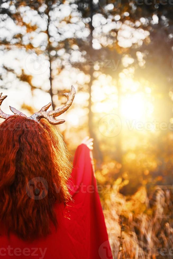 Mujer en vestido largo rojo con cuernos de ciervo en el bosque de otoño tratando de tocar una puesta de sol foto