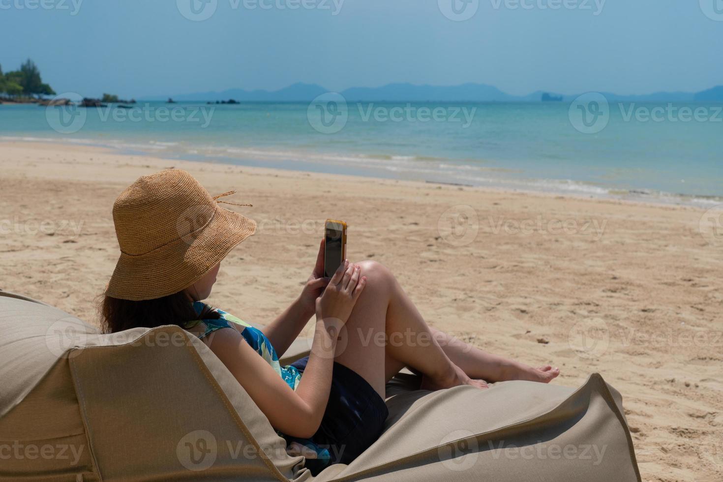 Mujer joven sentada en una bolsa de frijoles en la playa y con smartphone para tomar fotografías durante las vacaciones de verano foto