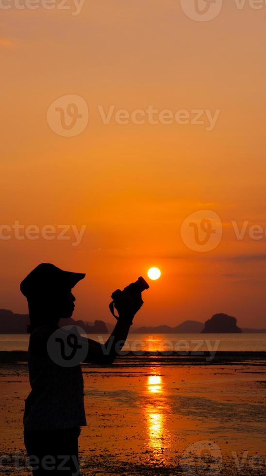 Silueta de mujer joven con cámara para tomar fotografías de la puesta de sol en la playa durante las vacaciones de verano foto