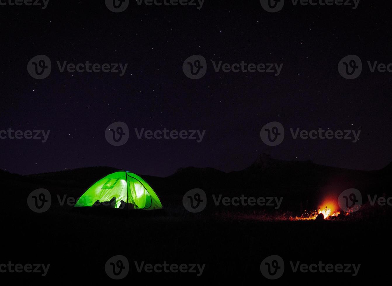 carpa verde iluminada de pie junto a la fogata en la naturaleza por la noche con fondo de noche estrellada. foto