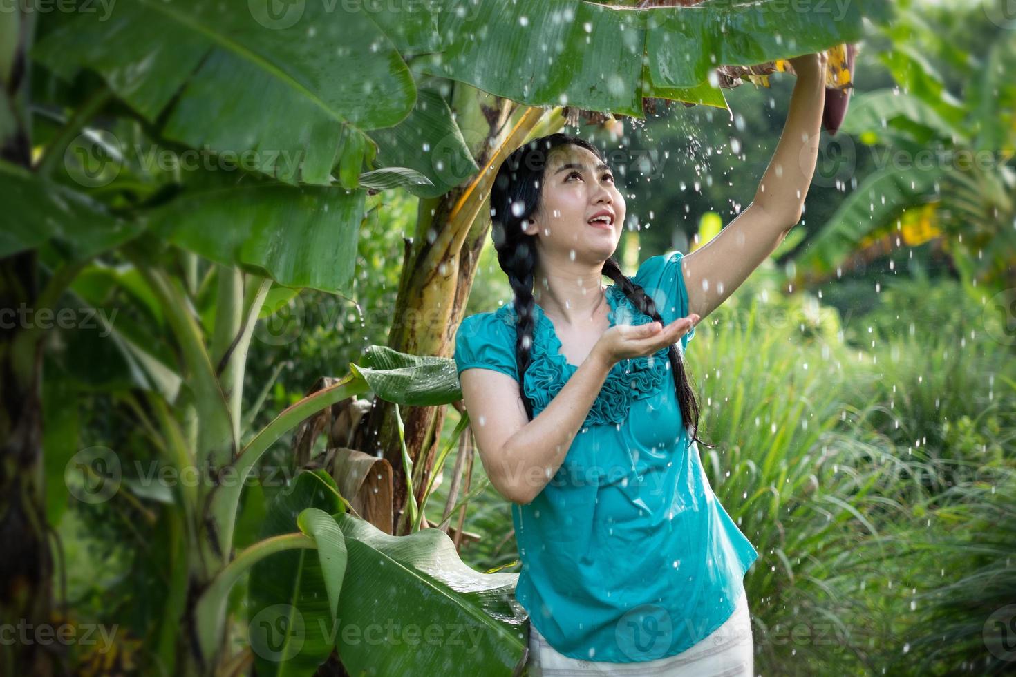 Retrato de una joven mujer asiática con cabello negro sosteniendo una hoja de plátano en la lluvia en el fondo del jardín verde foto