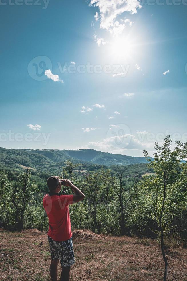 A man uses his binocular and looking over the trees and mountain hill.  A man in the red t-shirt looks at a beautiful landscape with binoculars. photo