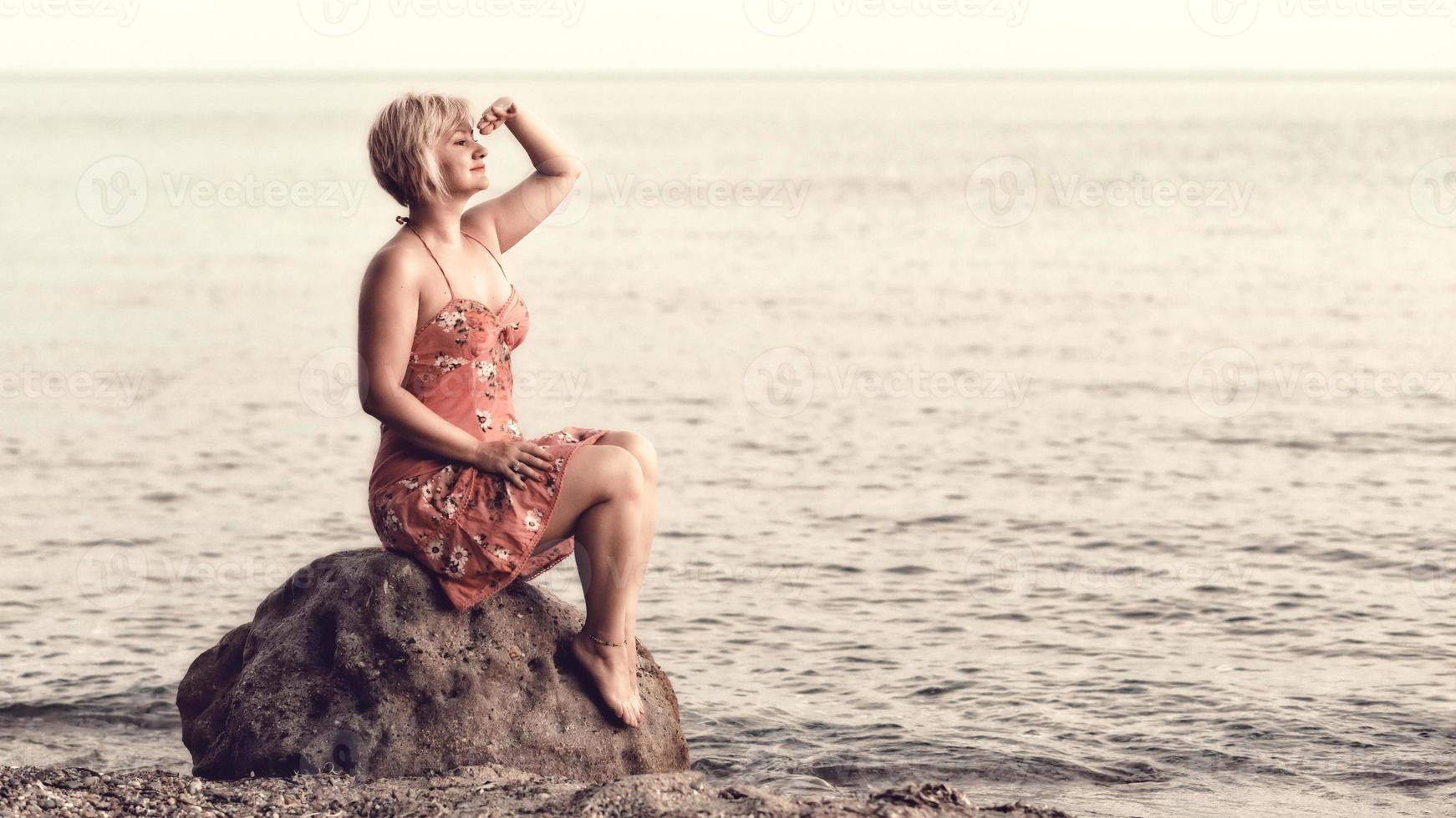 mujer rubia vestida de púrpura sentada en la enorme roca junto al mar, mirando la puesta de sol y esperando algo o alguien. foto