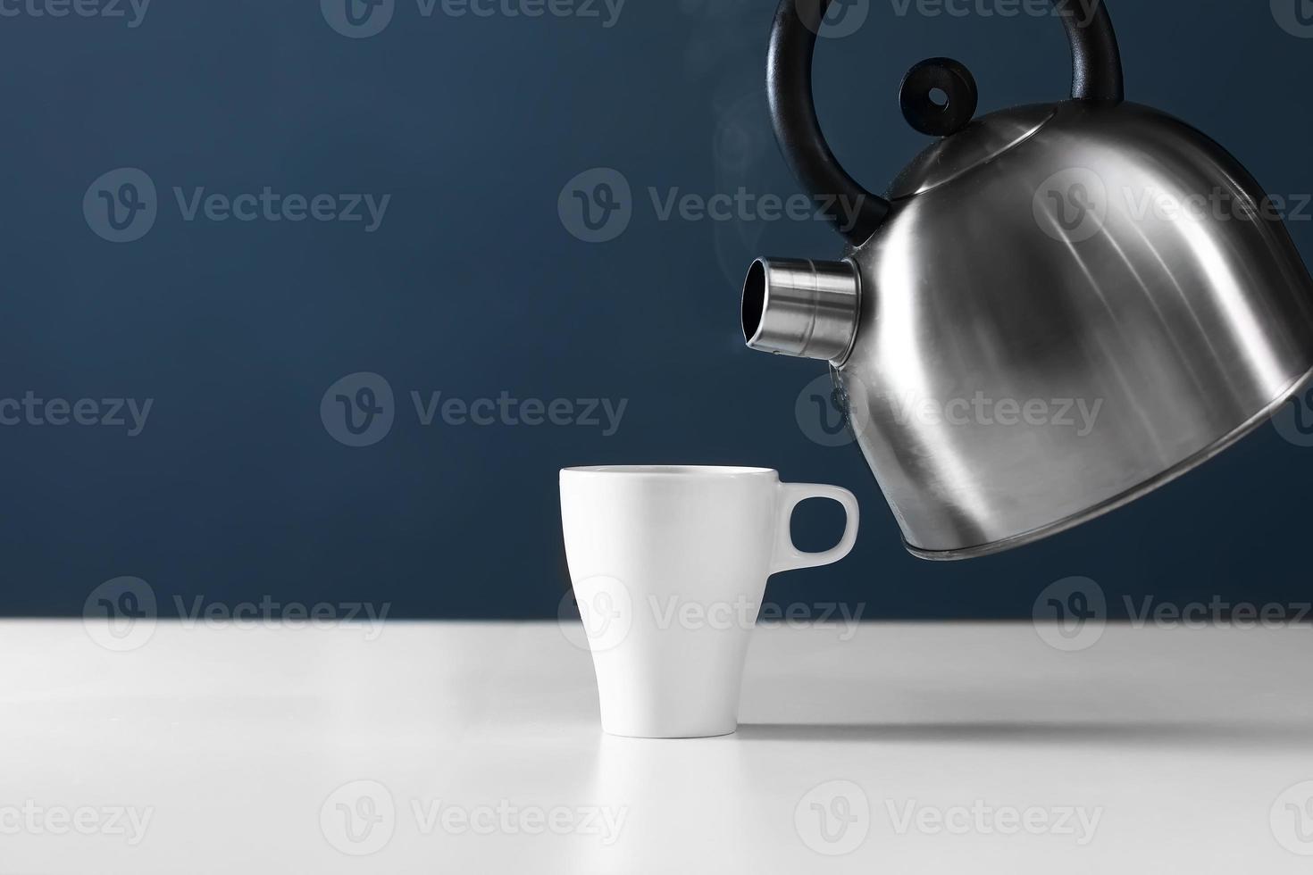 white mug with kettle on a white table. kettle pouring boiling water into a cup with smoke on wood table photo