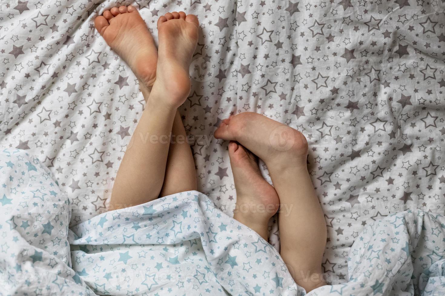 Close-up of two pairs of toddler girls feet on the bed under the blanket. Light blue and beige tones. photo