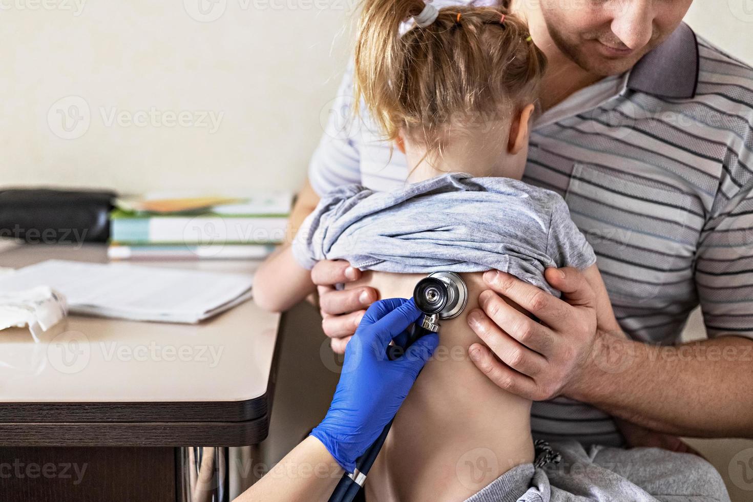 niña en los brazos de su padre en el consultorio del médico en la clínica. el médico examina al niño, escucha los pulmones con un fonendoscopio. tratamiento y prevención de infecciones respiratorias. foto