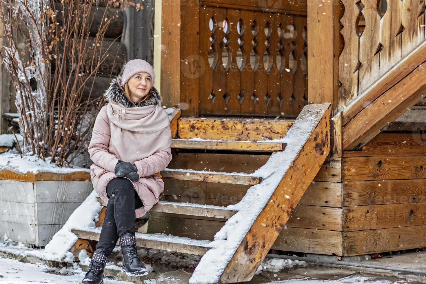 Mujer joven cálidamente vestida posando en el porche de una casa de madera en el pueblo. vacaciones de invierno en el campo foto