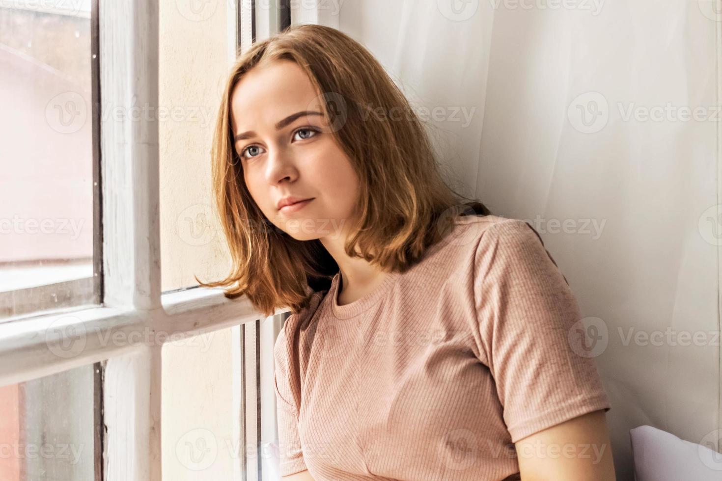 pensativa hermosa joven sentada en un alféizar cerca de la ventana.Retrato de un adolescente solo en casa.Concepto de personas, estilo de vida, ocio y relajación. foto