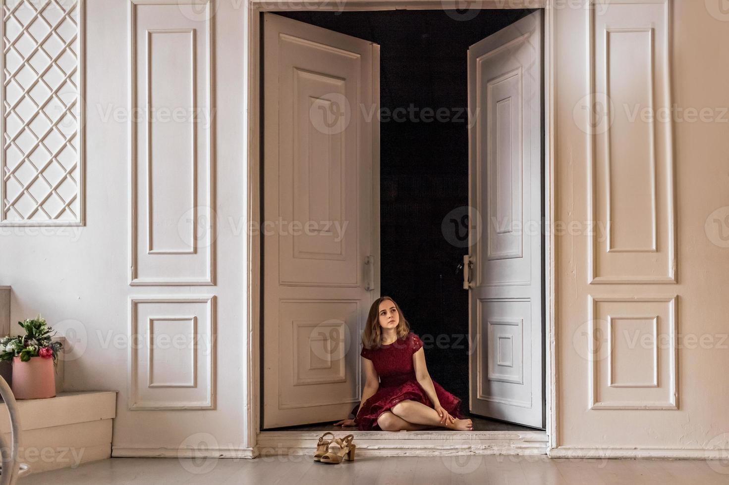 A cute stylish young girl in an elegant burgundy dress sits in the doorway to her room. Teenager. Graduation in school, college photo