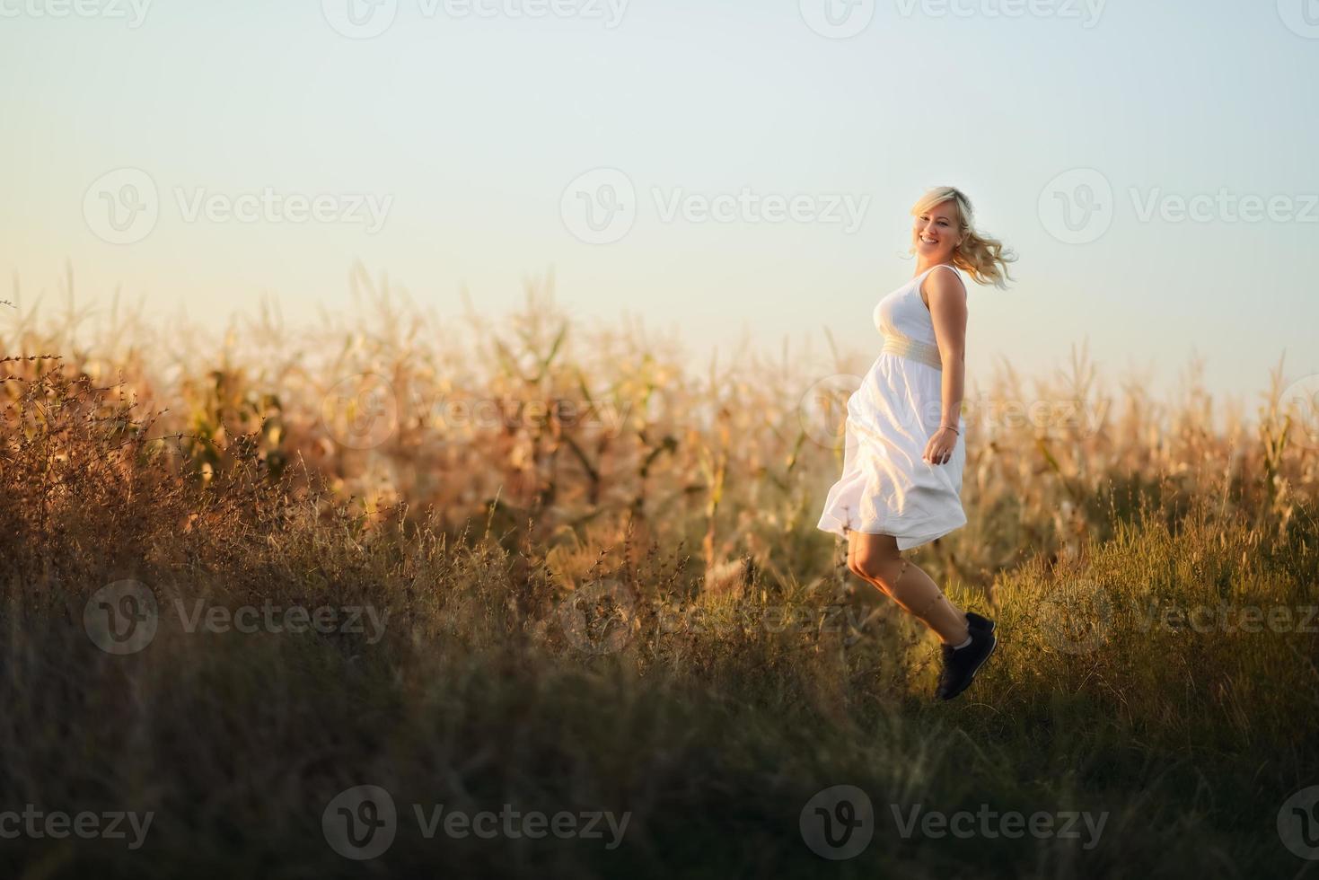 mujer feliz con un vestido largo de verano blanco saltando frente al campo de maíz. foto