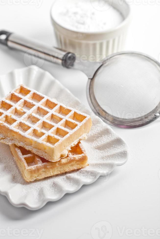 Obleas de Bélgica con azúcar en polvo en un plato de cerámica y un colador en la mesa blanca. foto