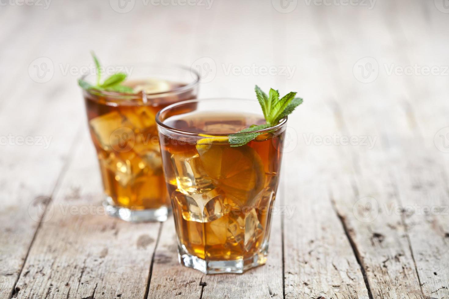 Dos vasos con té helado tradicional con limón, hojas de menta y cubitos de hielo en vidrio sobre fondo de mesa de madera rústica. foto