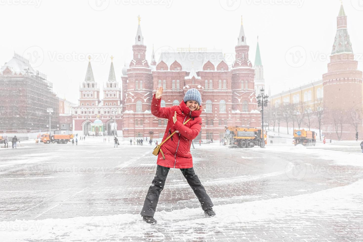 una hermosa joven con una chaqueta roja camina por la plaza manezhnaya en moscú durante una nevada y una ventisca. los quitanieves están trabajando en segundo plano. foto