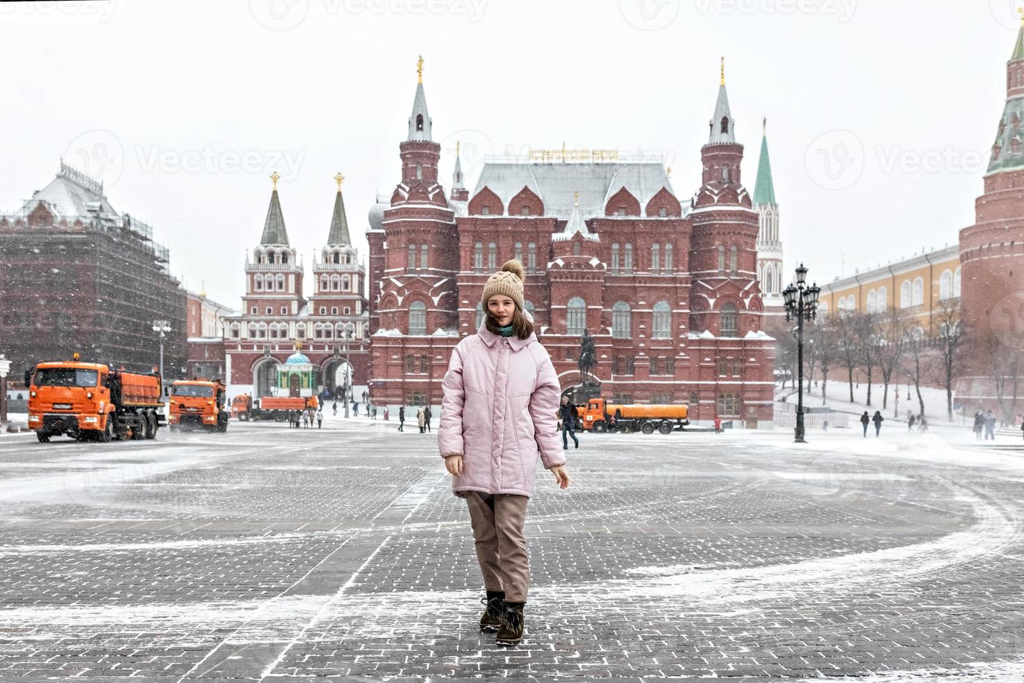 una hermosa joven con una chaqueta rosa camina por la plaza manezhnaya en moscú durante una nevada y una ventisca. los quitanieves están trabajando en segundo plano. foto