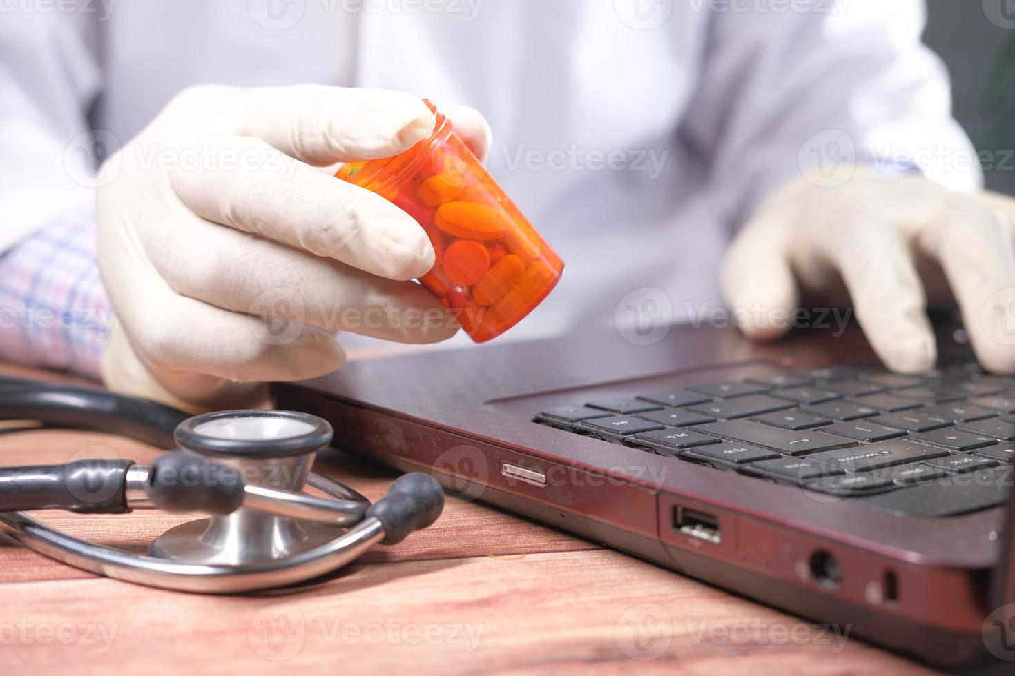 Mano en guantes de látex sosteniendo un contenedor de píldoras médicas y usando una computadora portátil foto