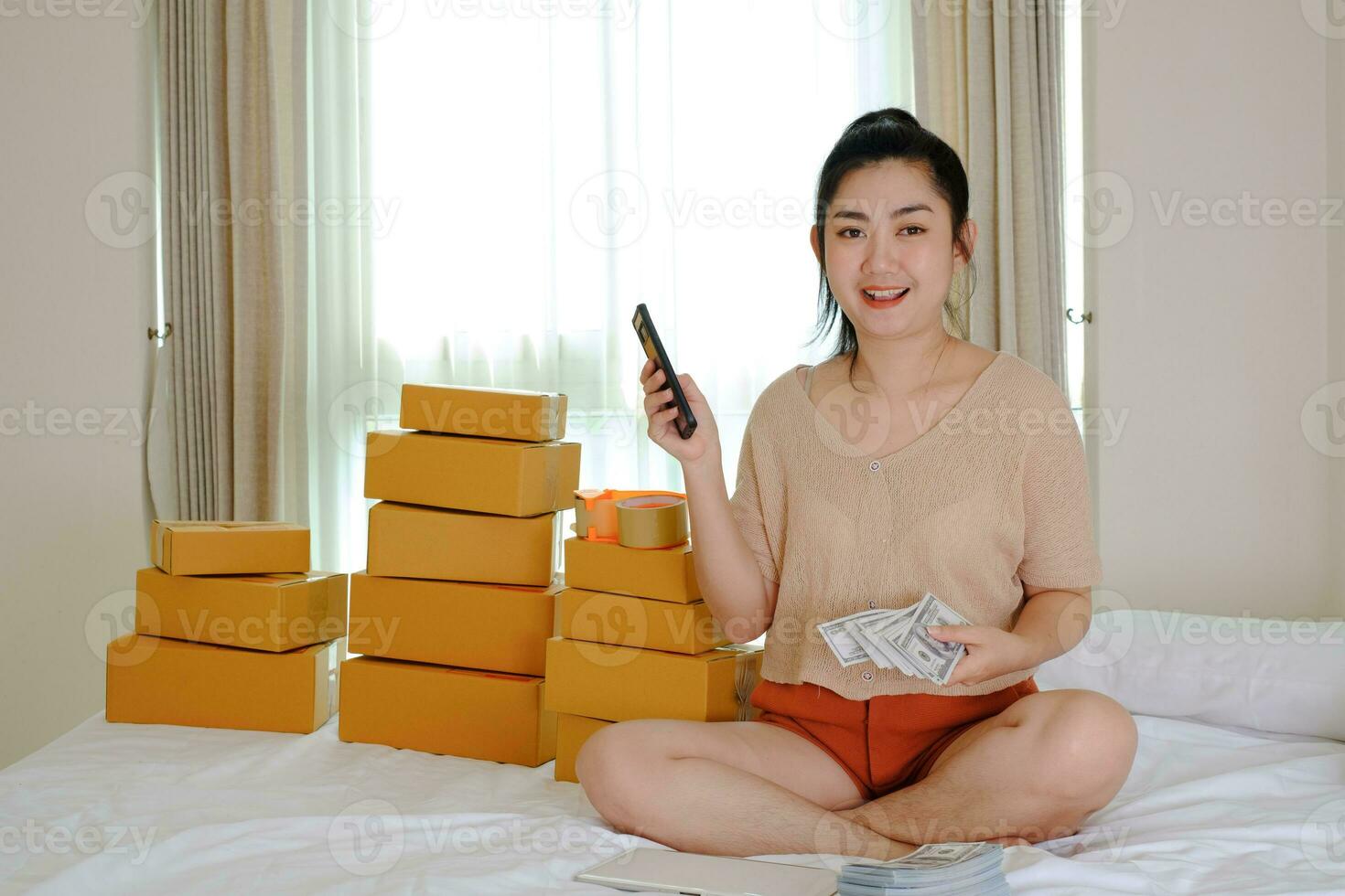 Portrait Asia young woman is sitting on the bed holding money paper currency 100 USD  phone with the phone at her bedroom photo