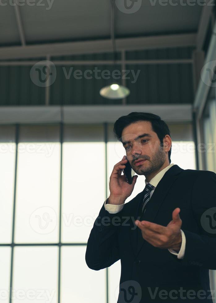 Candid portrait of young handsome caucasian confident businessman in black suit with tie, using smartphone to talk with client in office photo