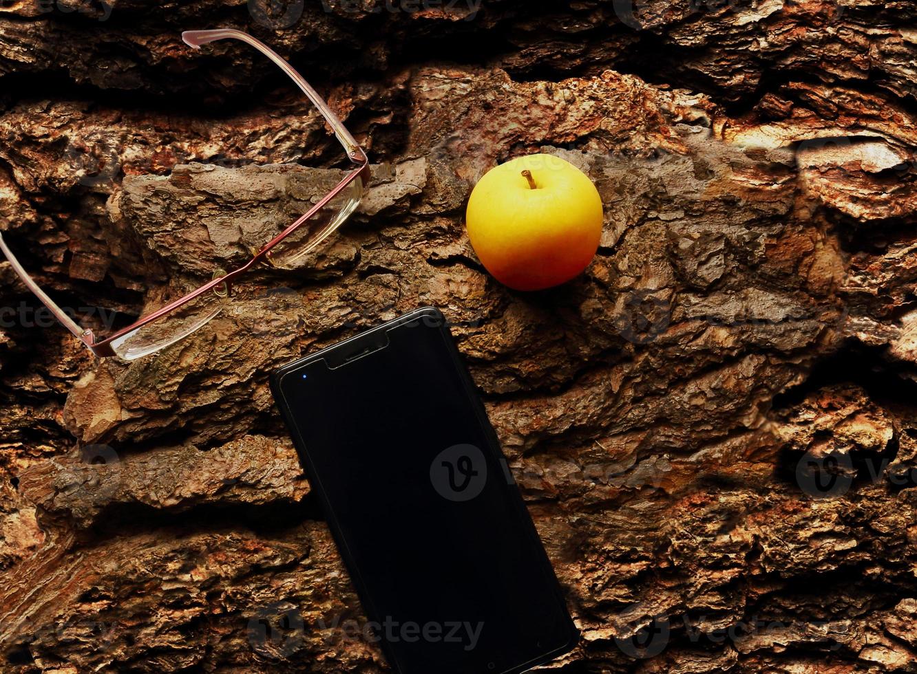 Yellow apple, glasses and cellphone flat lay on tree bark photo