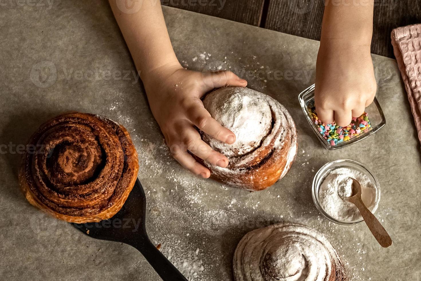 Children's hands in the frame sprinkle a fresh baked bun with cinnamon photo