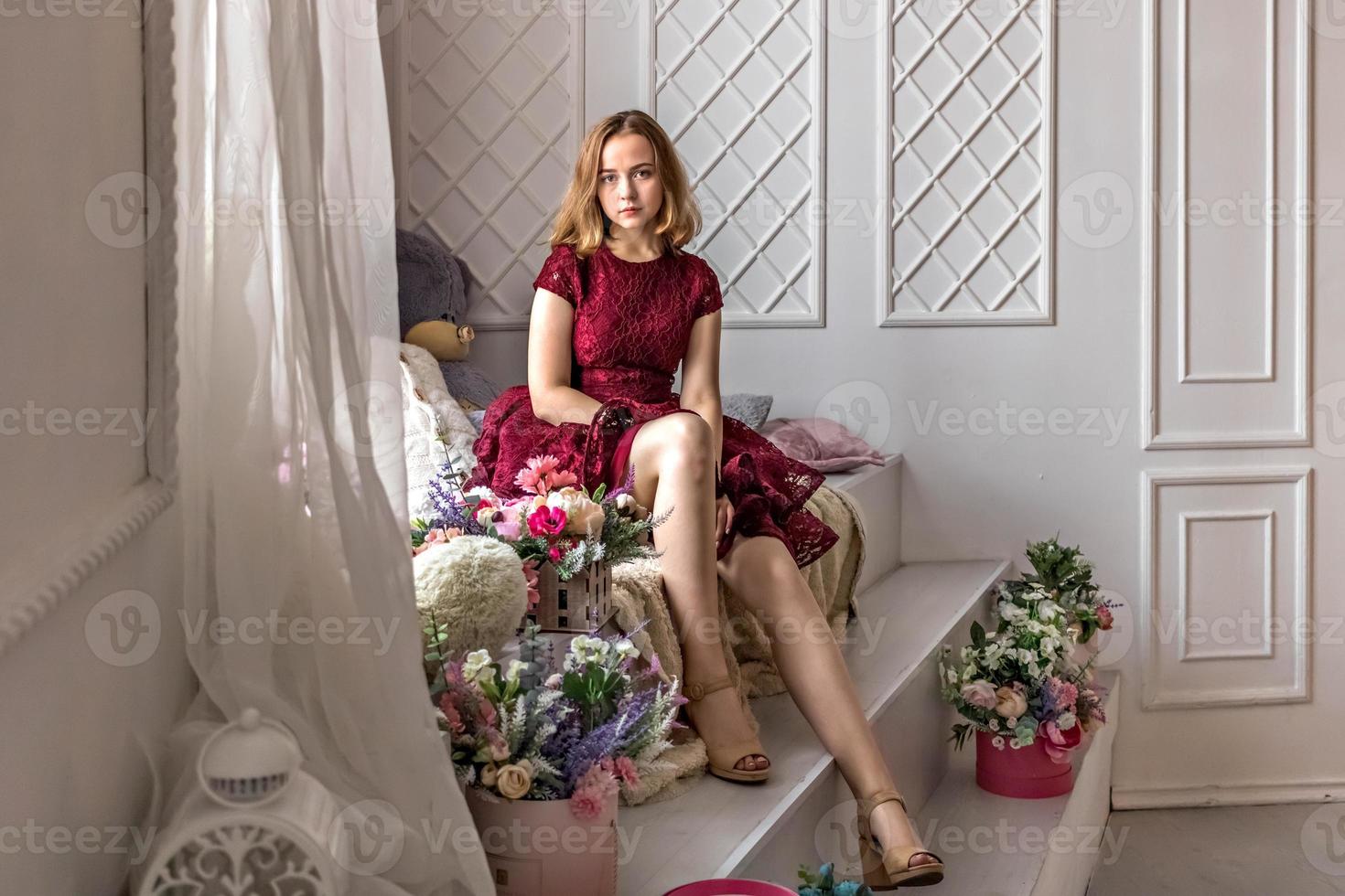 A cute stylish young girl in an elegant burgundy dress sits by the window photo