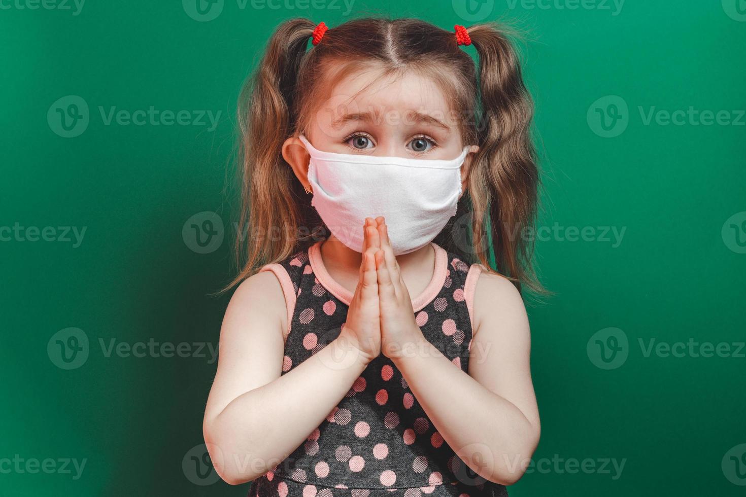 Caucasian sick little girl in medical mask during coronavirus epidemic prays on green background closeup photo