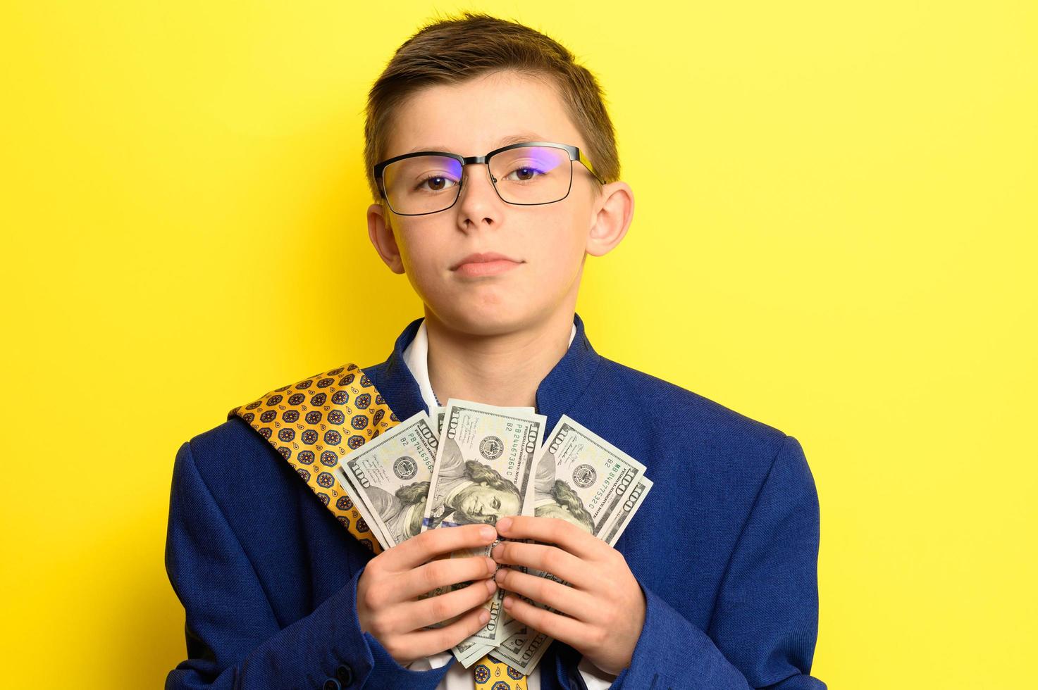 A boy in glasses and an oversized suit with a satisfied face is holding US dollars, a child and big money. photo