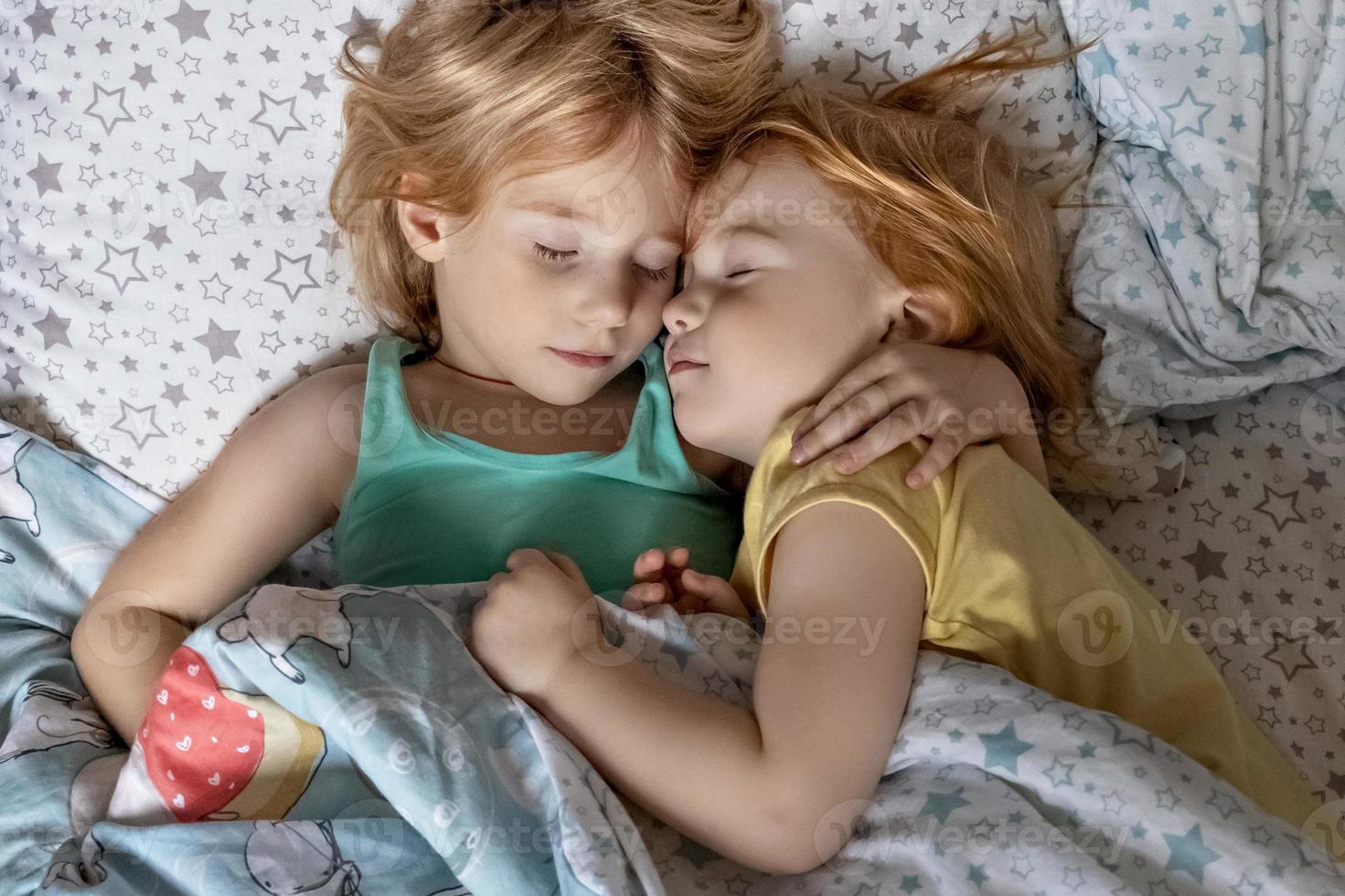 Two little sibling girls sisters sleeping in an embrace in bed under one blanket photo