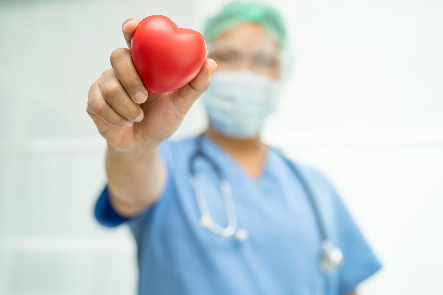 Asian doctor wearing face shield and PPE suit new normal to protect safety infection Covid-19 Coronavirus holding red heart in hand at hospital. photo