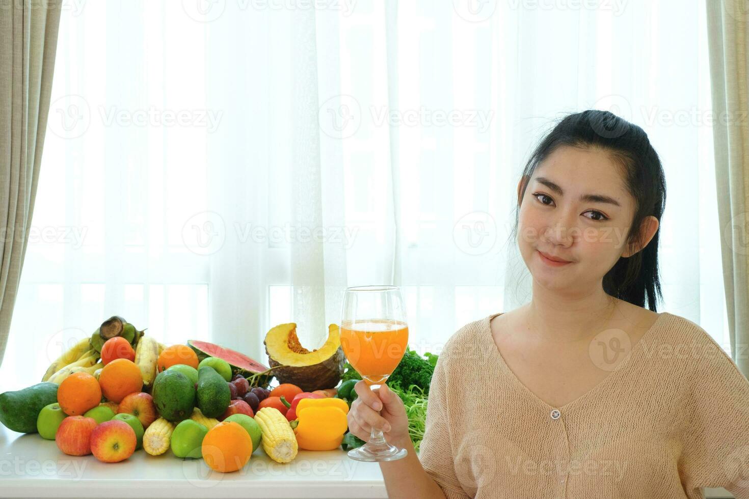 Retrato mujer mano sosteniendo jugo de naranja recién exprimido en vaso con una variedad de frutas y verduras maduras frescas sobre la mesa en el fondo de la cortina blanca foto