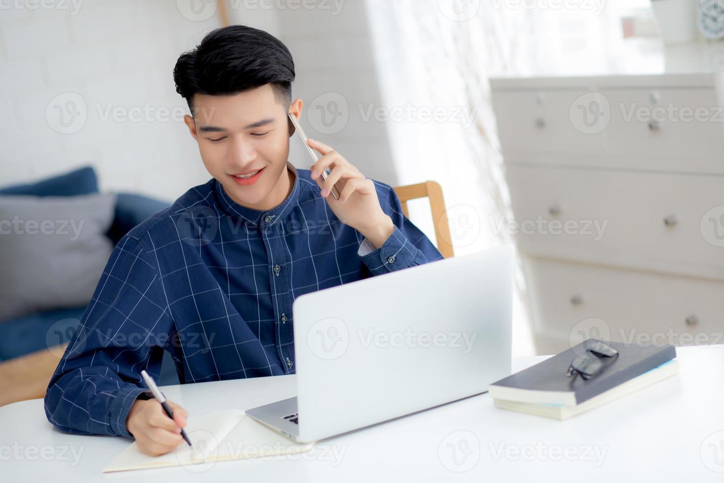 Hombre asiático joven que habla por teléfono y trabaja desde casa con una computadora portátil, un hombre independiente que escribe en un cuaderno y habla en un teléfono inteligente, se queda en casa, la vida doméstica, los negocios y el concepto de comunicación. foto