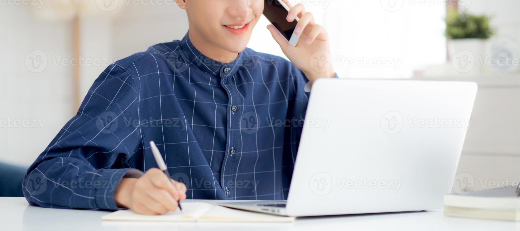 Young asian man talking phone and work from home with laptop computer, freelance male writing on notebook and speak on smartphone, stay home, domestic life, business and communication concept. photo