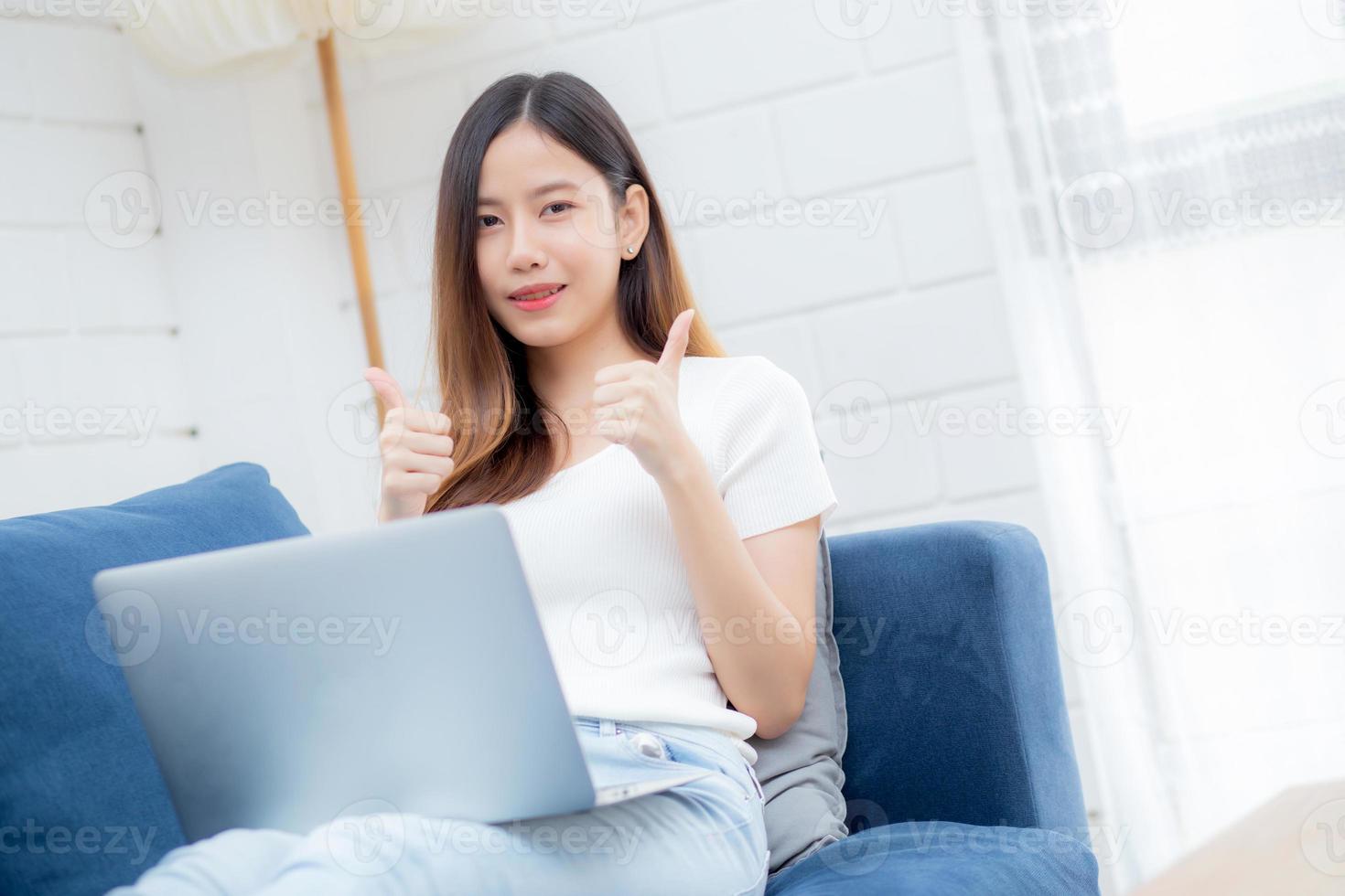 Joven mujer de negocios asiática sonríe y trabaja desde casa con una computadora portátil en línea a Internet en el sofá en la sala de estar, una chica independiente que usa un cuaderno en el sofá con comodidad, un nuevo concepto de estilo de vida normal. foto