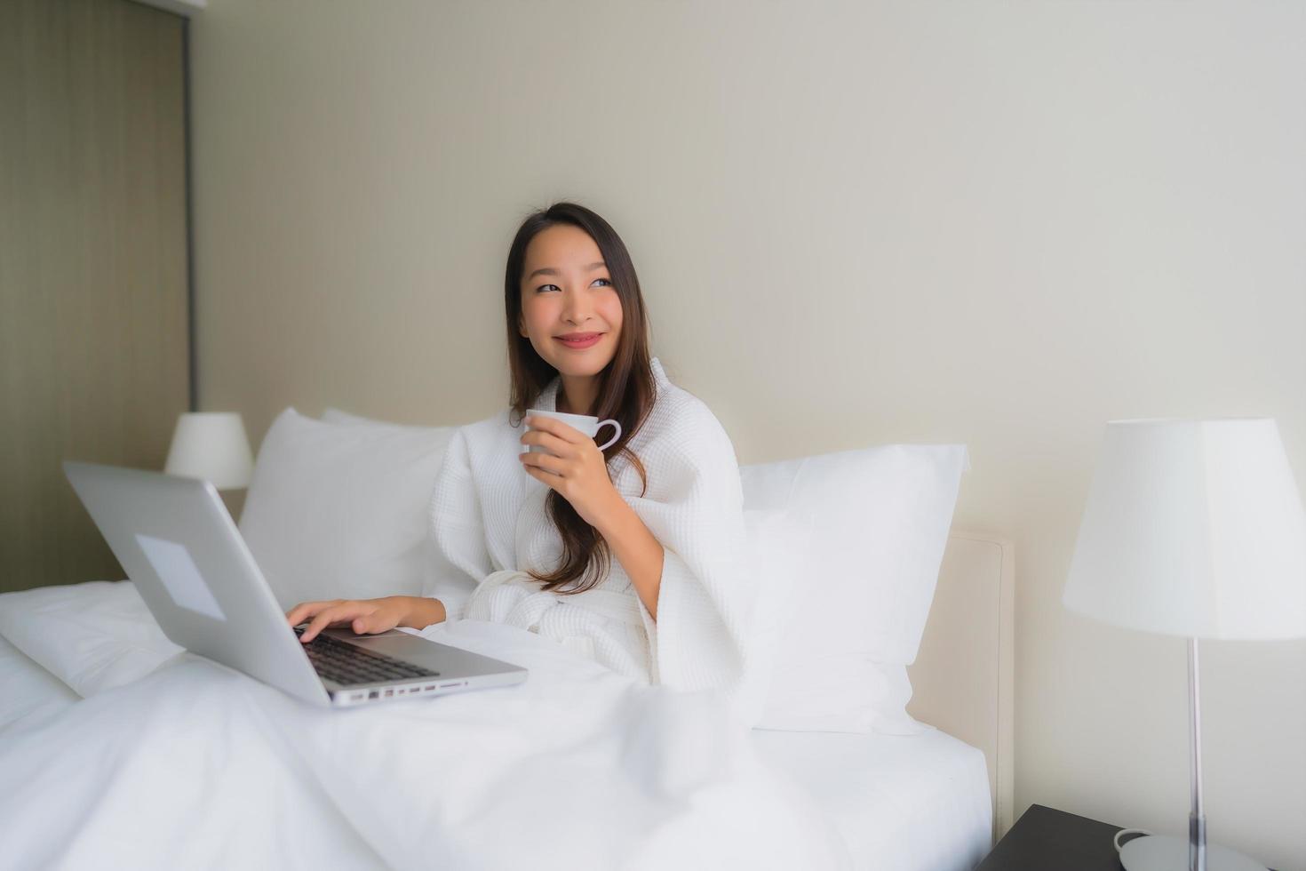 Retrato de hermosas mujeres asiáticas jóvenes con taza de café y computadora portátil en la cama foto