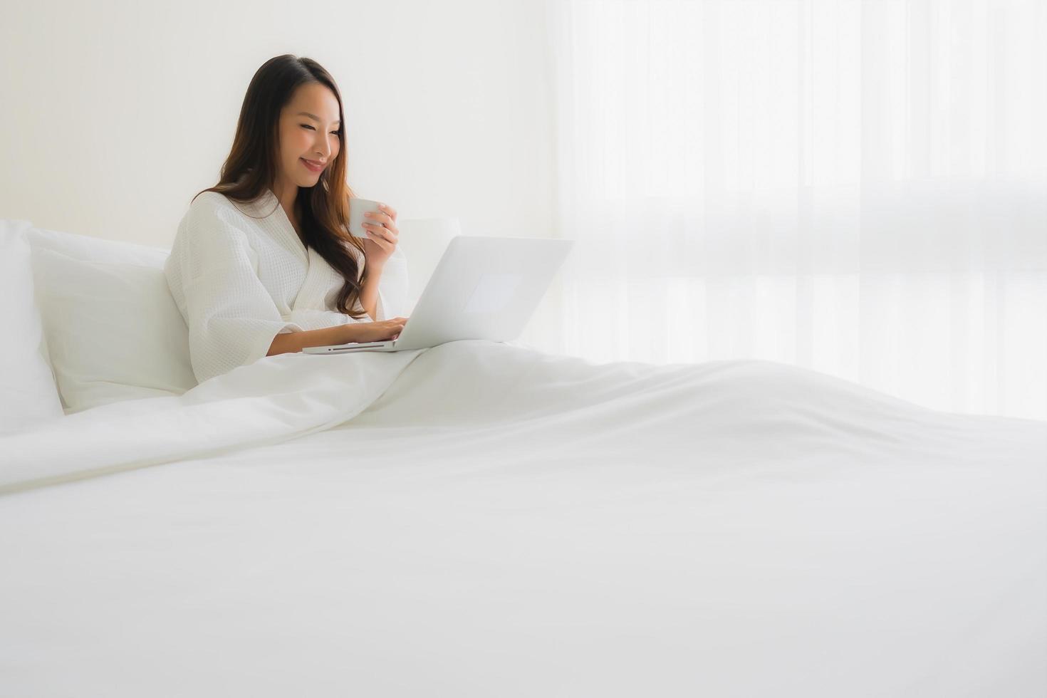 Retrato de hermosas mujeres asiáticas jóvenes con taza de café y computadora portátil en la cama foto