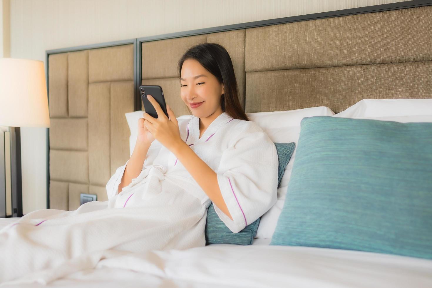 retrato, hermoso, joven, mujeres asiáticas, utilizar, móvil, teléfono inteligente, con, taza de café, en cama foto