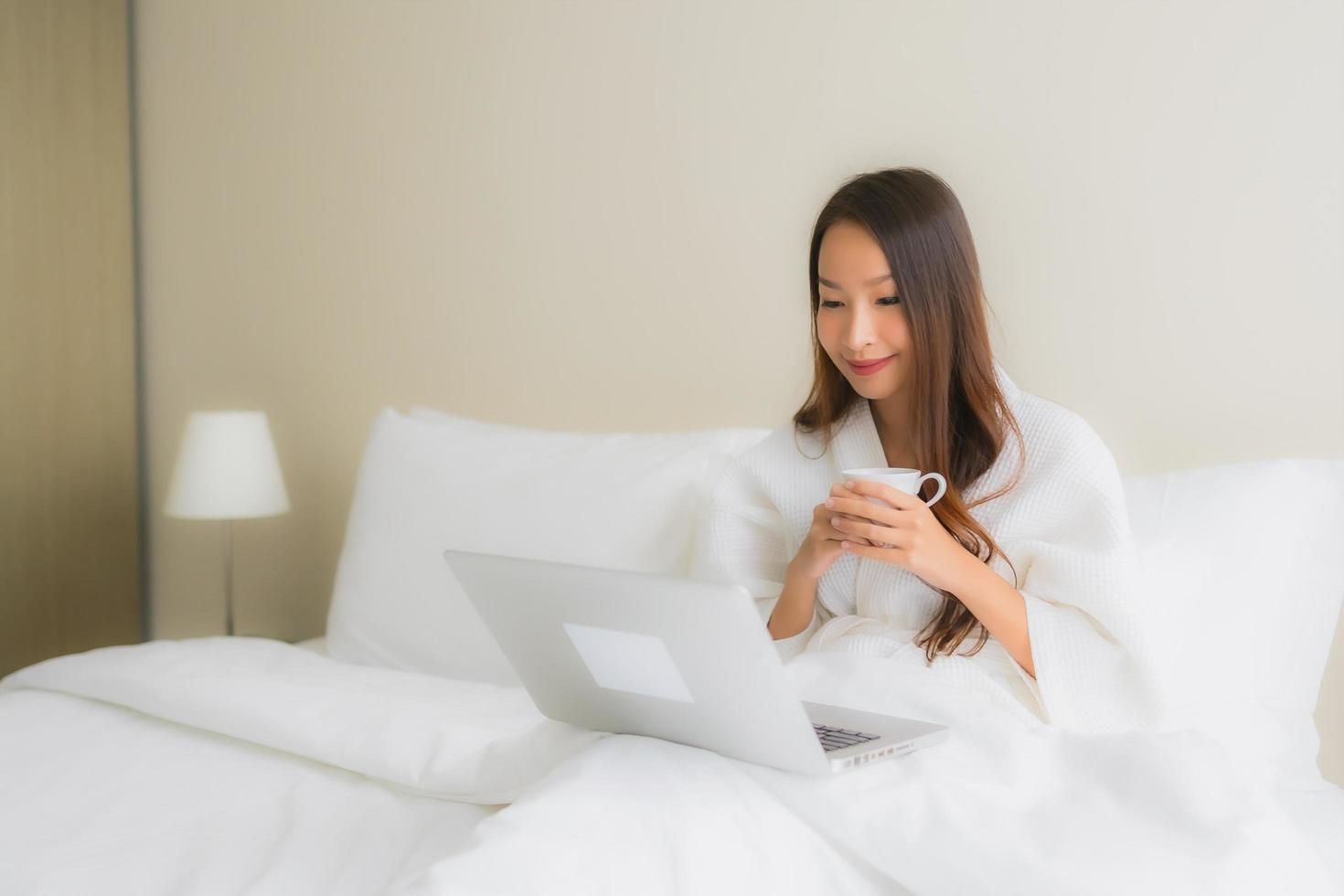 Retrato de hermosas mujeres asiáticas jóvenes con taza de café y computadora portátil en la cama foto