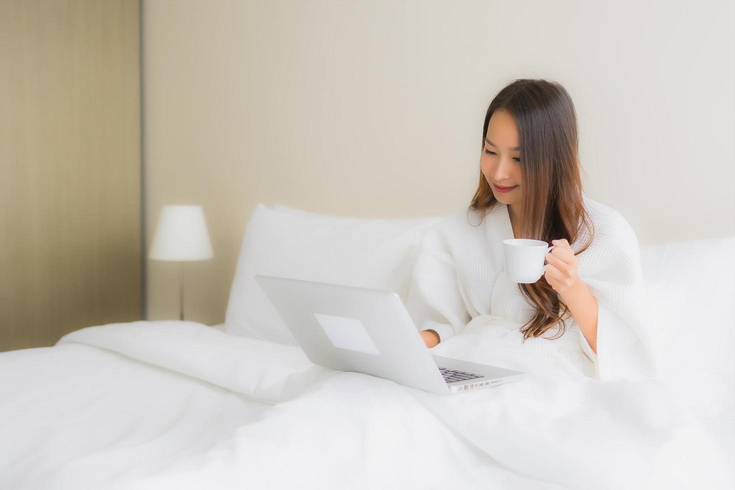 Retrato de hermosas mujeres asiáticas jóvenes con taza de café y computadora portátil en la cama foto