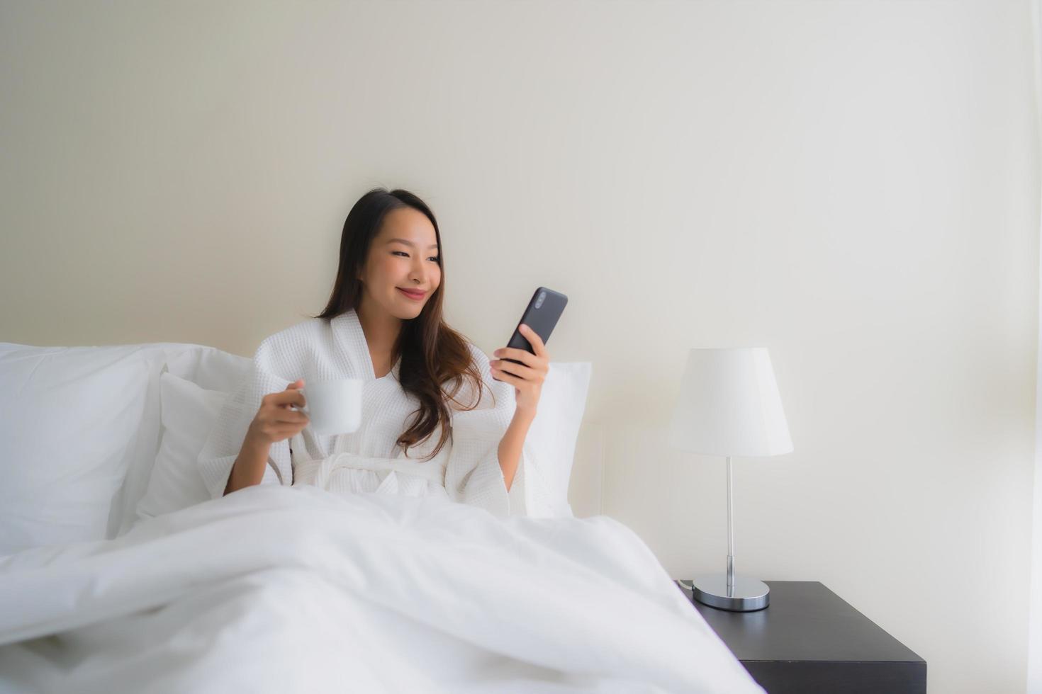 retrato, hermoso, joven, mujeres asiáticas, con, taza de café, y, teléfono móvil, en cama foto