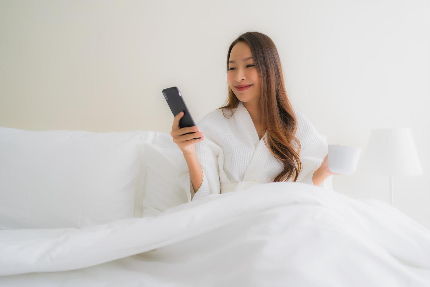 retrato, hermoso, joven, mujeres asiáticas, con, taza de café, y, teléfono móvil, en cama foto