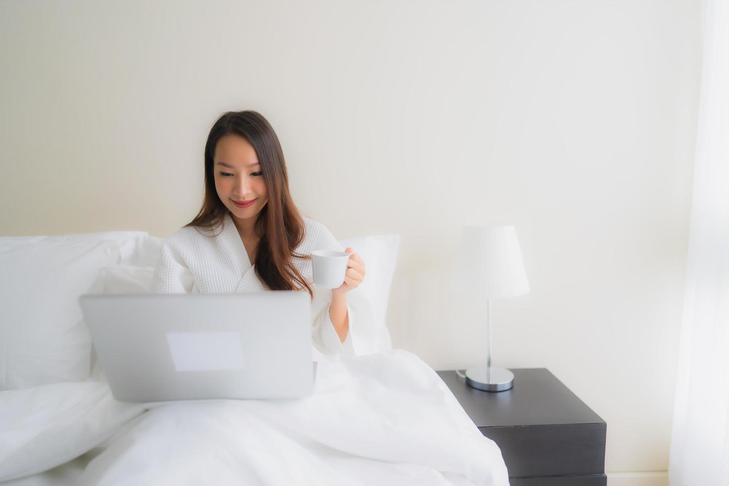 Retrato de hermosas mujeres asiáticas jóvenes con taza de café y computadora portátil en la cama foto