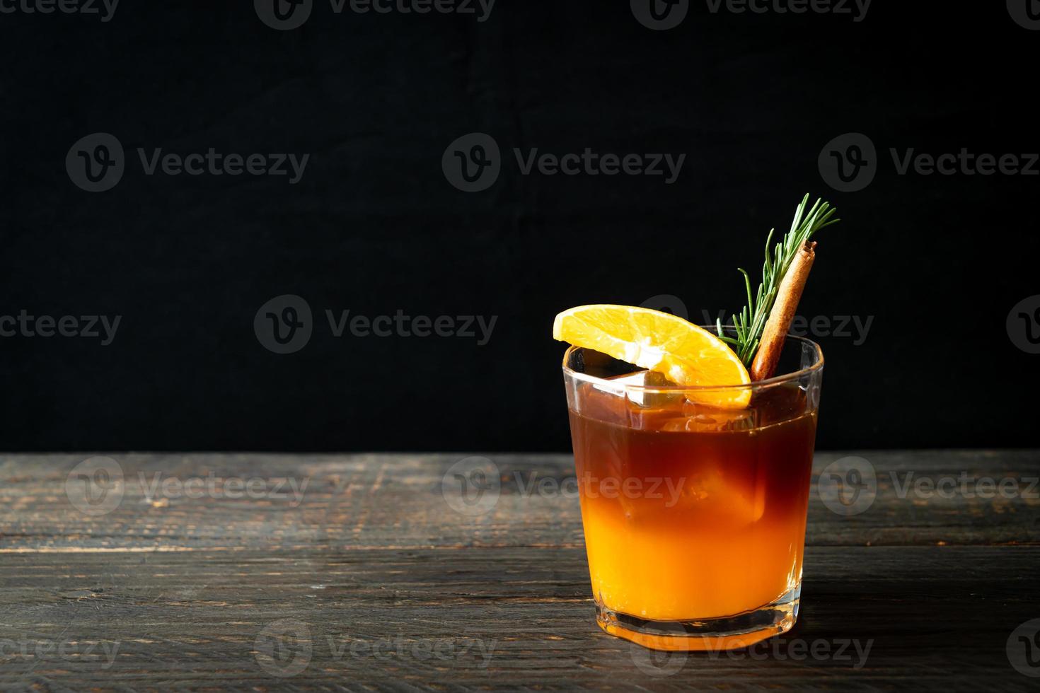 A glass of iced Americano black coffee and a layer of orange and lemon juice decorated with rosemary and cinnamon on a wood background photo