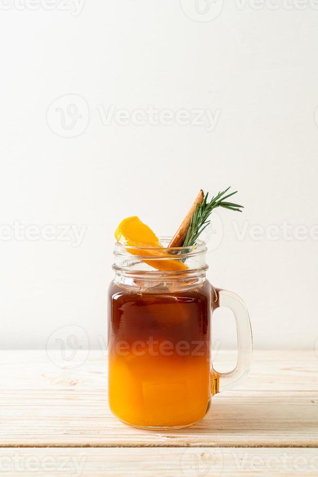 A glass of iced Americano black coffee and a layer of orange and lemon juice decorated with rosemary and cinnamon on a wood background photo