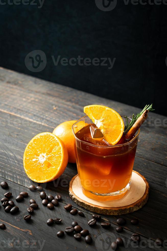 A glass of iced Americano black coffee and a layer of orange and lemon juice decorated with rosemary and cinnamon on a wood background photo