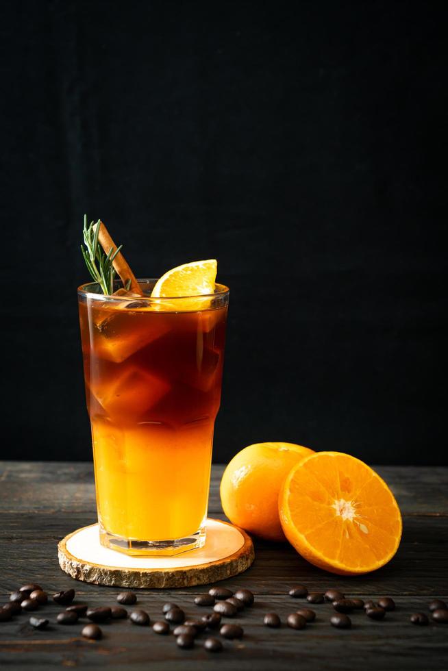 A glass of iced Americano black coffee and a layer of orange and lemon juice decorated with rosemary and cinnamon on a wood background photo