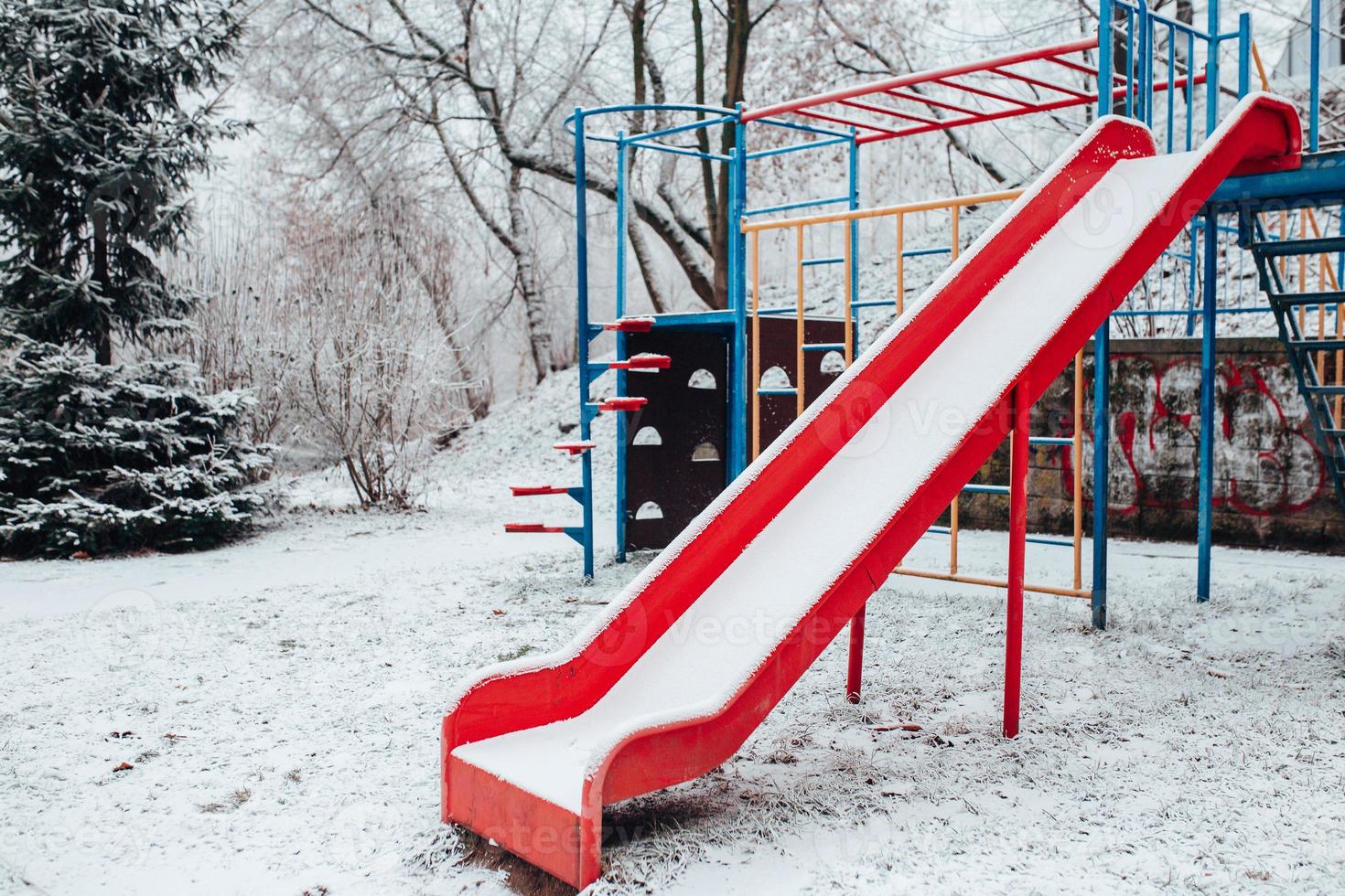 Columpio para bebés cubierto de nieve en invierno - parque infantil vacío - columpio de plástico rojo en el frío foto