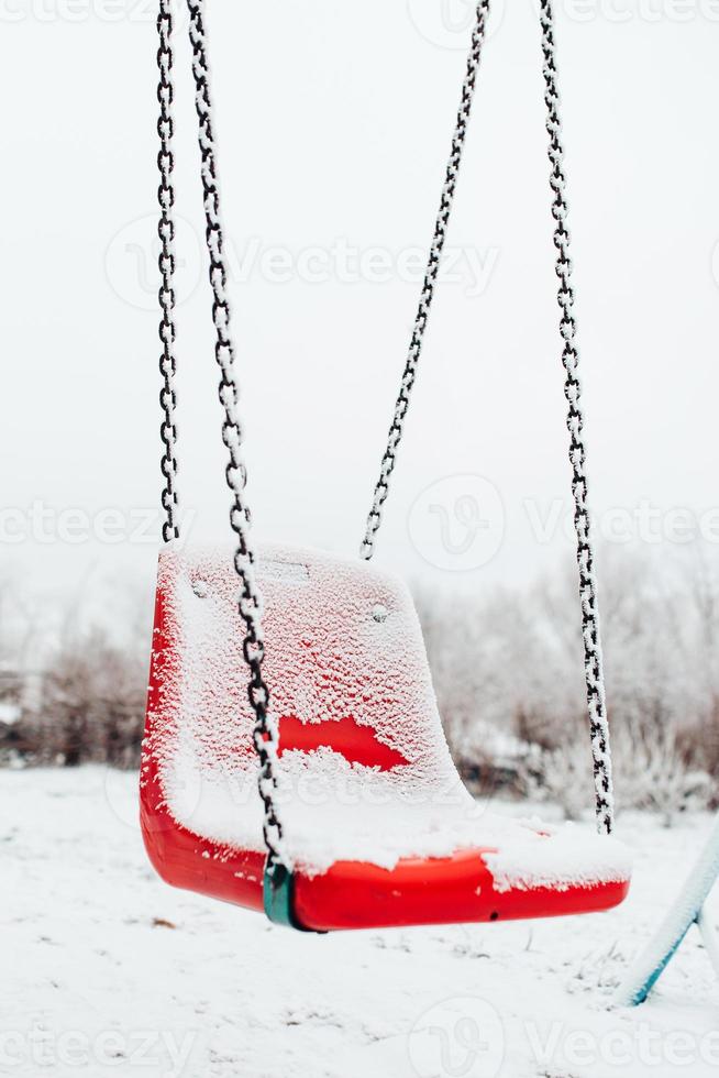 Columpio para bebés cubierto de nieve en invierno - parque infantil vacío - columpio de plástico rojo en el frío foto