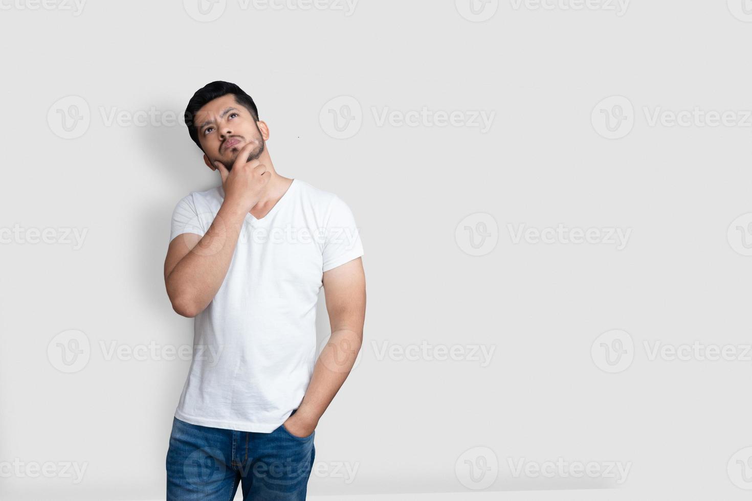 Hombre guapo asiático en camiseta blanca que tiene dudas mientras mira hacia arriba sobre fondo blanco aislado foto