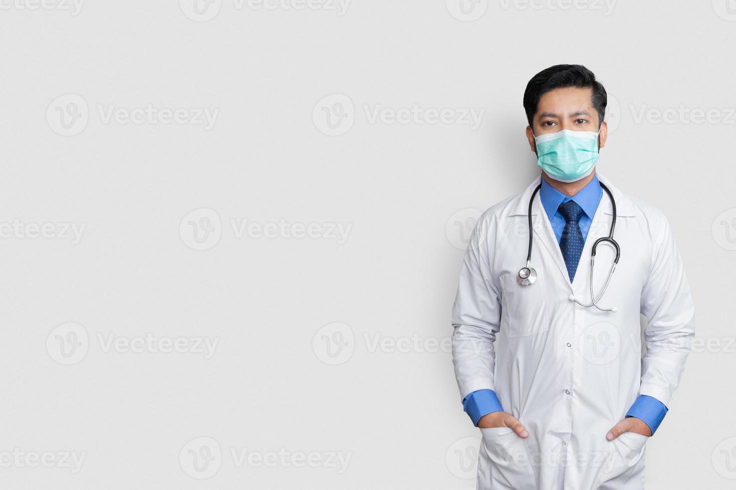 Médico varón joven rostro cubierto con máscara y las manos en el bolsillo del abrigo sobre antecedentes aislados, concepto de salud foto