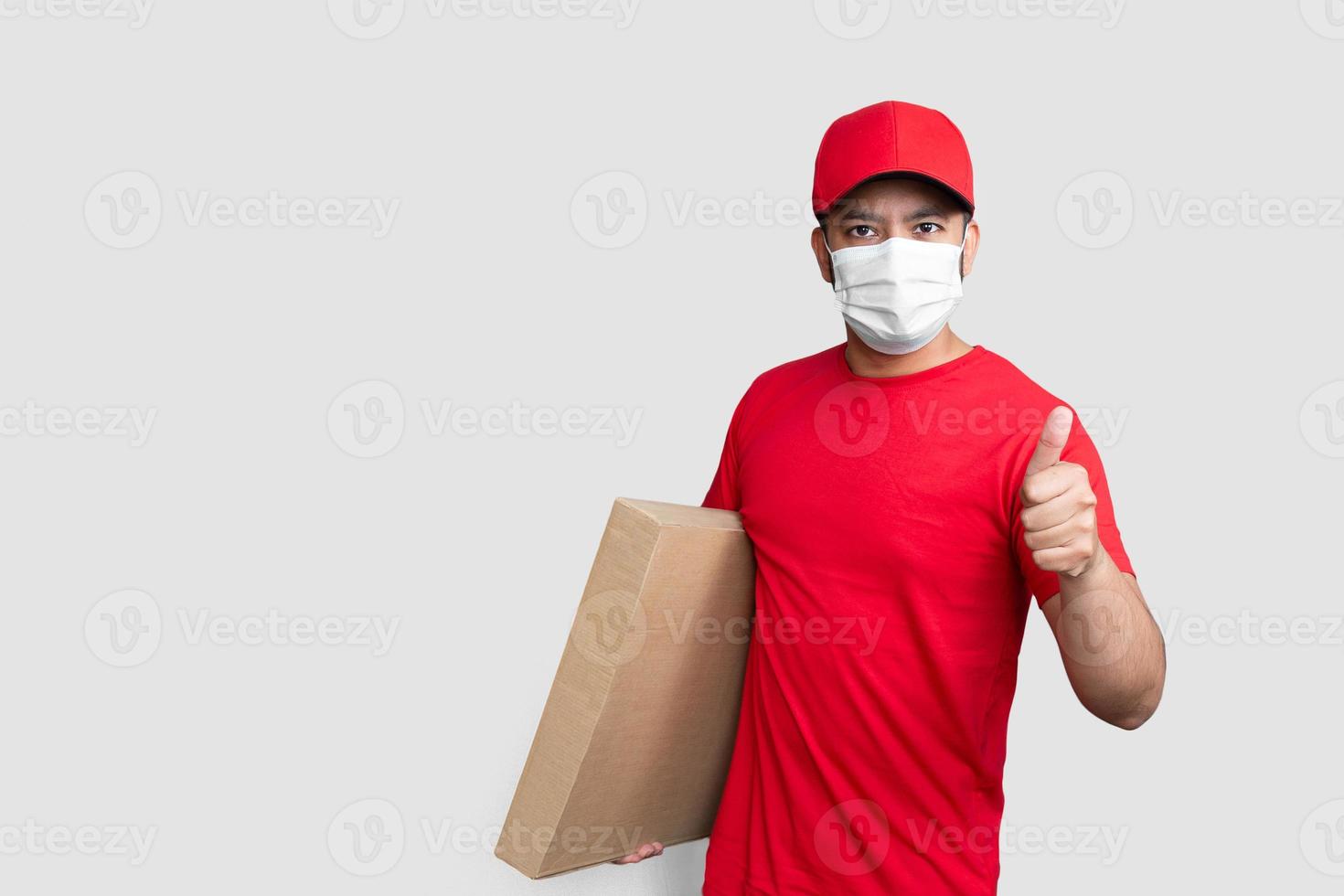 Repartidor empleado en gorra roja camiseta en blanco uniforme mascarilla mantenga la caja de cartón vacía mostrando los pulgares hacia arriba aislado sobre fondo blanco. foto