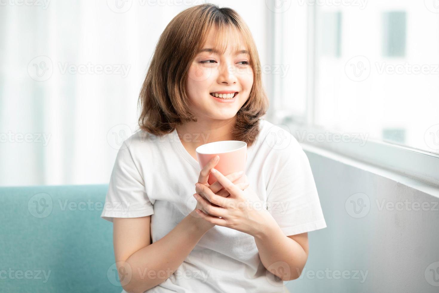 Young girl holding cup sitting by glass door in early morning photo